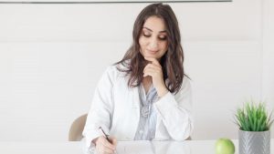 A female nutritionist in a lab coat writing notes with an apple and plant on the desk.