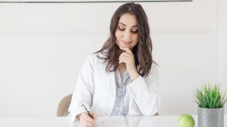 A female nutritionist in a lab coat writing notes with an apple and plant on the desk.